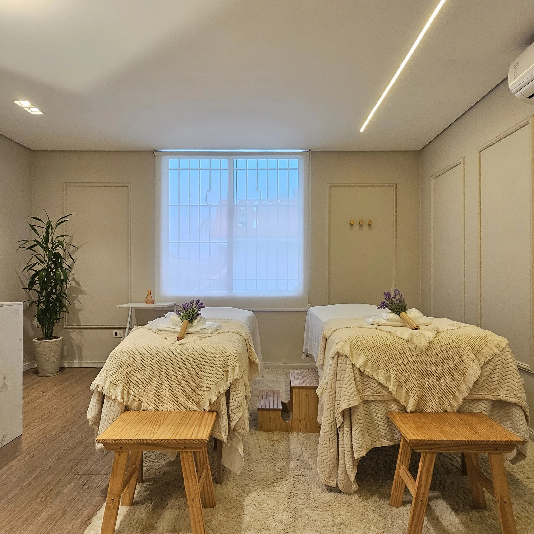 Sala de massagem com duas macas, mantas de tricô, ramos de lavanda e luz de janela